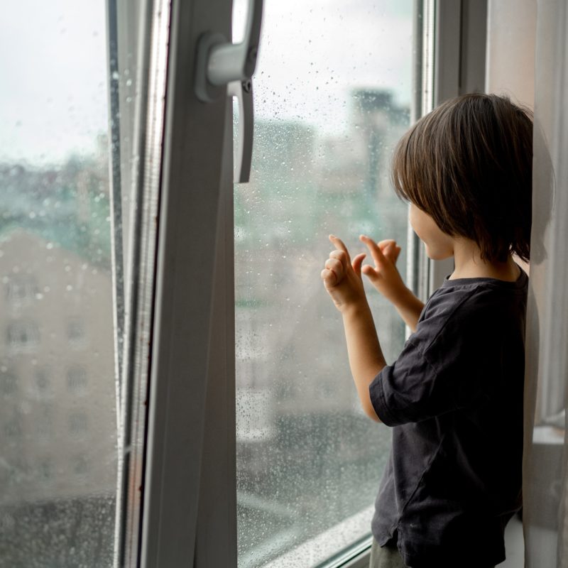 The baby is standing at the window on the windowsill.